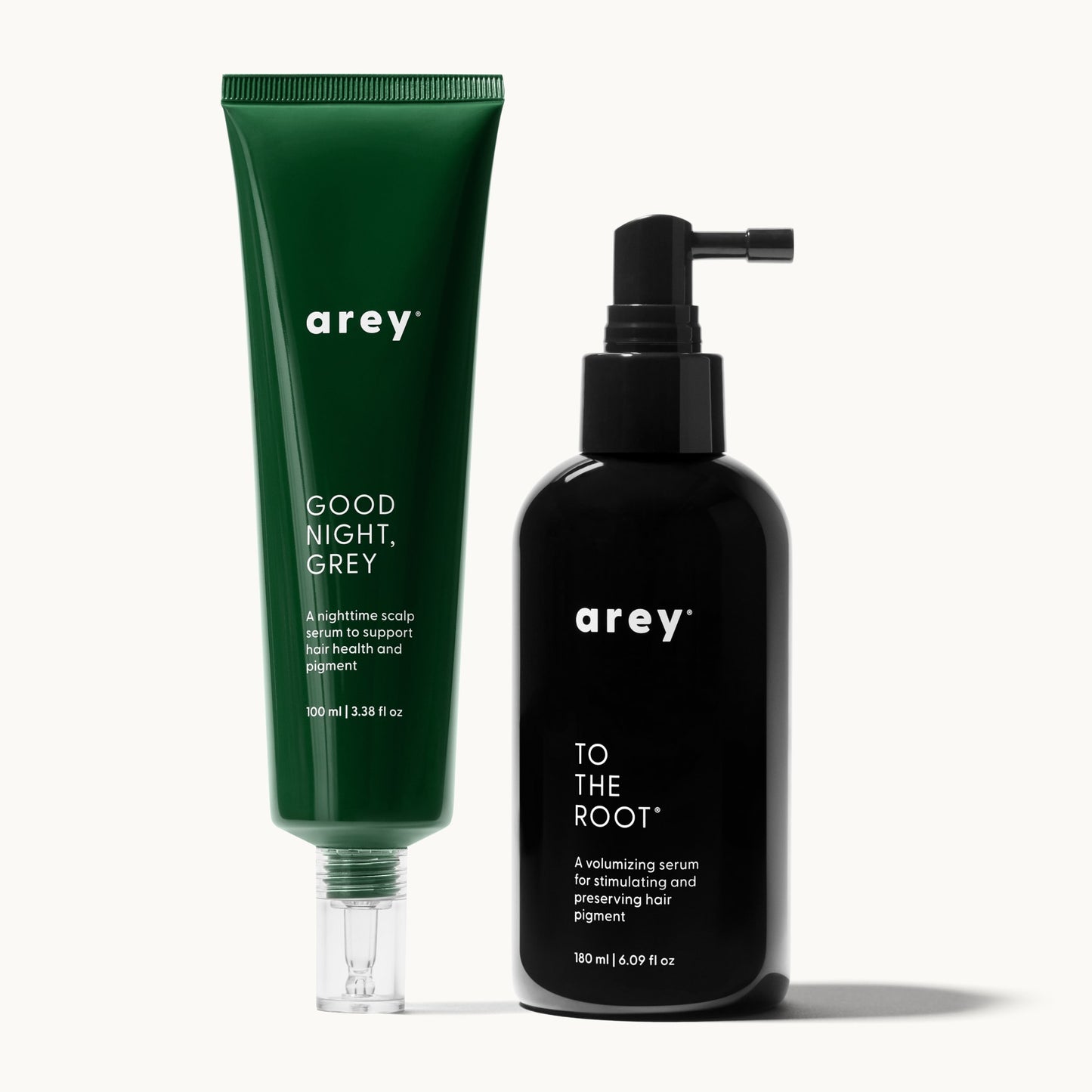Two Arey hair serum products on a cream background. On the left, a tall green tube labeled "arey® GOOD NIGHT, GREY – A nighttime scalp serum to support hair health and pigment – 100 ml | 3.38 fl oz." On the right, a black pump bottle labeled "arey® TO THE ROOT® – A volumizing serum for stimulating and preserving hair pigment – 180 ml | 6.09 fl oz."