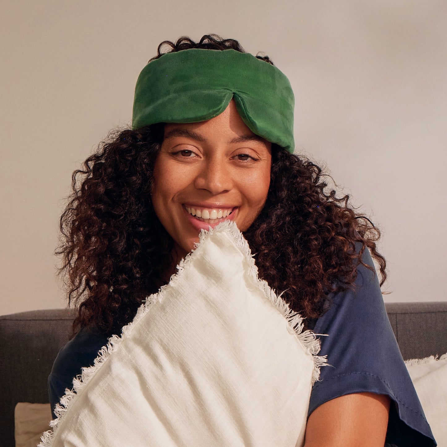 A smiling woman with curly hair, embodying the essence of hair wellness, sits on a couch holding a white pillow while wearing the Arey Beauty Sleep Duo’s green silk sleep mask on her forehead.