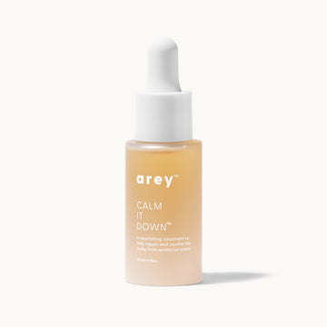 Product photo of the Arey Calm It Down™ scalp serum on a cream background. Golden glass dropper bottle (29ml) with white dropper cap, described as "a nourishing treatment to help repair and soothe the scalp from oxidative stress."