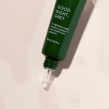 A green bottle labeled Good Night, Grey® by Arey, featuring a dropper tip, rests on a light surface with clear liquid drops nearby. This overnight scalp treatment is specially formulated to support hair health and pigment, featuring hair growth peptides designed to promote fuller, more vibrant hair.