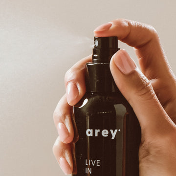 A hand holds a black spray bottle labeled “Arey Live In Mist Leave-In Conditioner,” pressing the nozzle to release a fine mist. The neutral background emphasizes the product and hand, showcasing its UV protection benefits and nourishment from Raspberry Seed Oil.