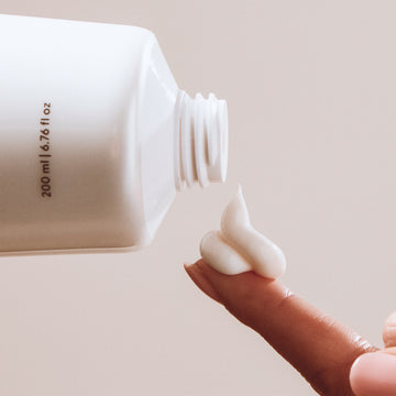 Hand holding a small amount of white conditioner next to a larger white container.