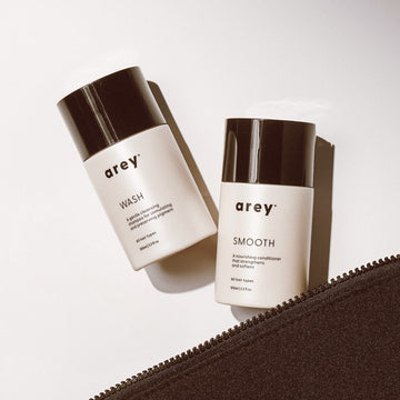 Two Arey Mini Duo bottles, labeled WASH and SMOOTH, rest on a white surface beside a partially open black zipper pouch. Soft lighting creates gentle shadows, accentuating the minimal and modern design of these science-forward Arey hair care essentials made for healthier hair.