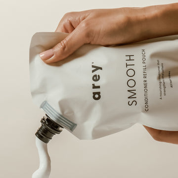 A hand squeezes Arey Smooth Conditioner Refill from a white pouch, dispensing white cream into an unseen container—a nourishing conditioner designed to repair and care for hair.