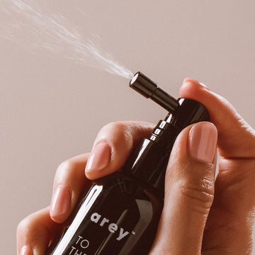 A hand with neatly manicured nails holds a black spray bottle of Arey’s To The Root® Scalp Serum, dispensing a fine mist designed to repigment greys and support aging hair.
