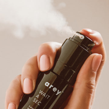 A hand with light pink nails holds a black spray bottle labeled Arey Wait A Sec® Dry Shampoo, releasing a fine, non-aerosol mist into the air against a neutral background; this dry shampoo by Arey is designed to absorb oil and boost volume.