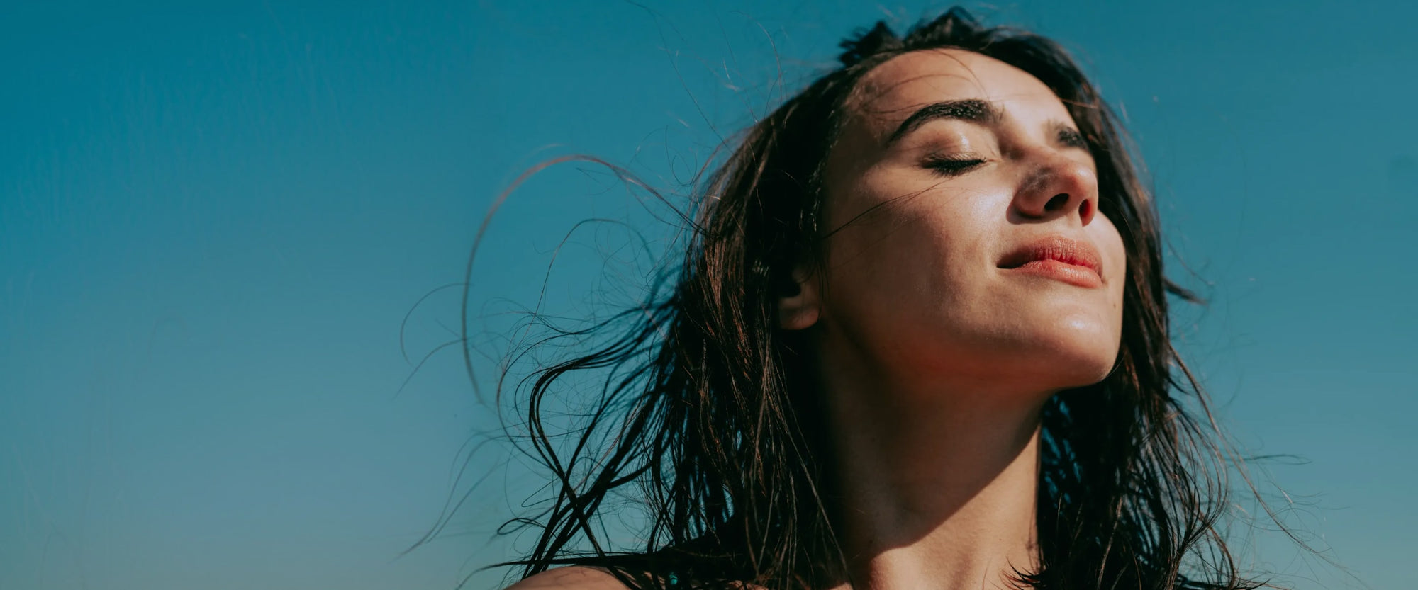 A person with long dark hair stands with eyes closed, facing the sun and sky, enjoying the warmth and breeze.