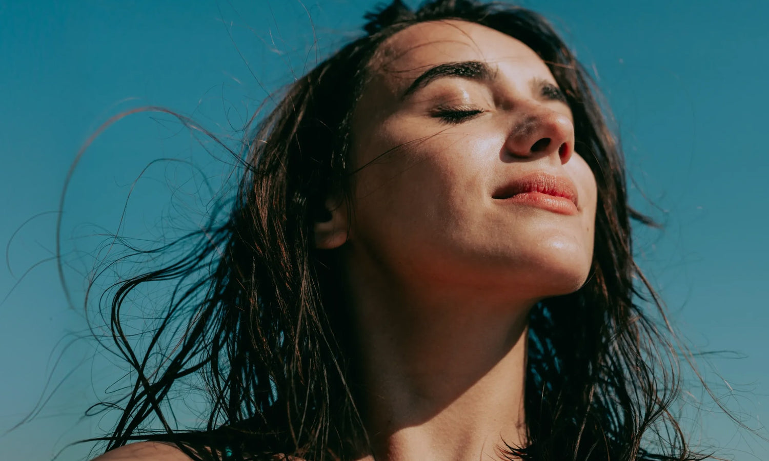 A woman with long dark hair stands with her eyes closed and face turned toward the sun, enjoying the warmth. Blue sky is in the background and her hair is gently windswept.