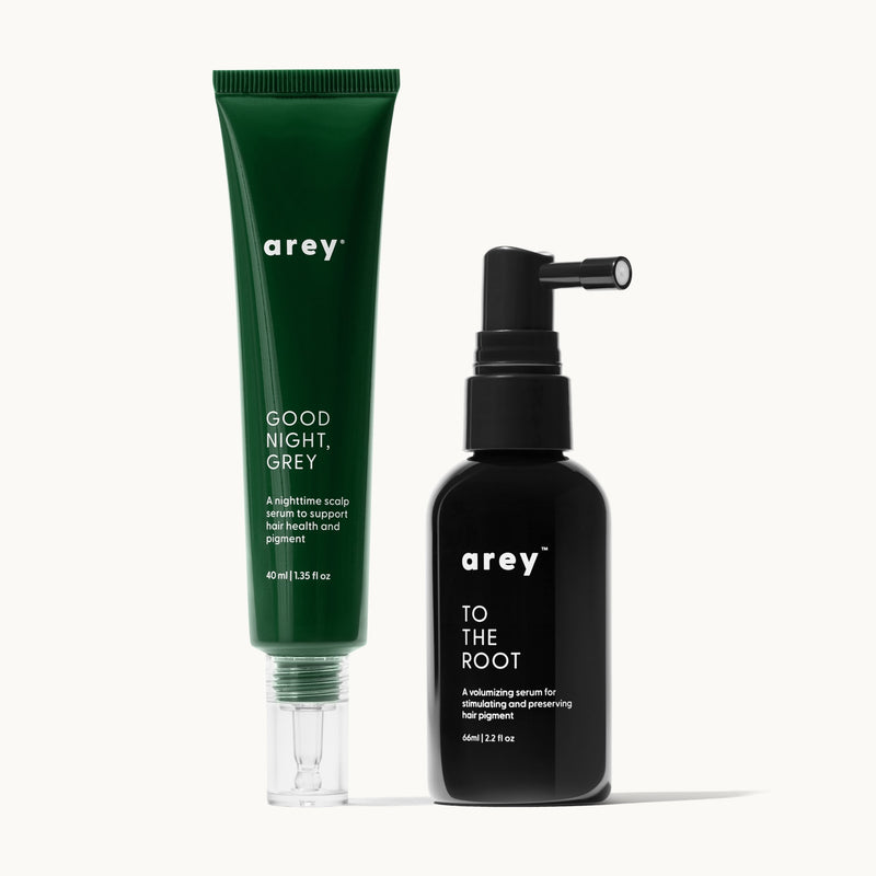 Two Arey hair serum products on a cream background. On the left, a green tube labeled "arey® GOOD NIGHT, GREY – A nighttime scalp serum to support hair health and pigment – 40 ml | 1.35 fl oz." On the right, a black spray bottle labeled "arey™ TO THE ROOT – A volumizing serum for stimulating and preserving hair pigment – 66ml | 2.2 fl oz."