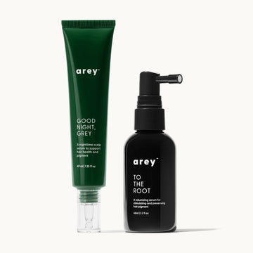 Two Arey hair serum products on a cream background. On the left, a green tube labeled "arey® GOOD NIGHT, GREY – A nighttime scalp serum to support hair health and pigment – 40 ml | 1.35 fl oz." On the right, a black spray bottle labeled "arey™ TO THE ROOT – A volumizing serum for stimulating and preserving hair pigment – 66ml | 2.2 fl oz."
