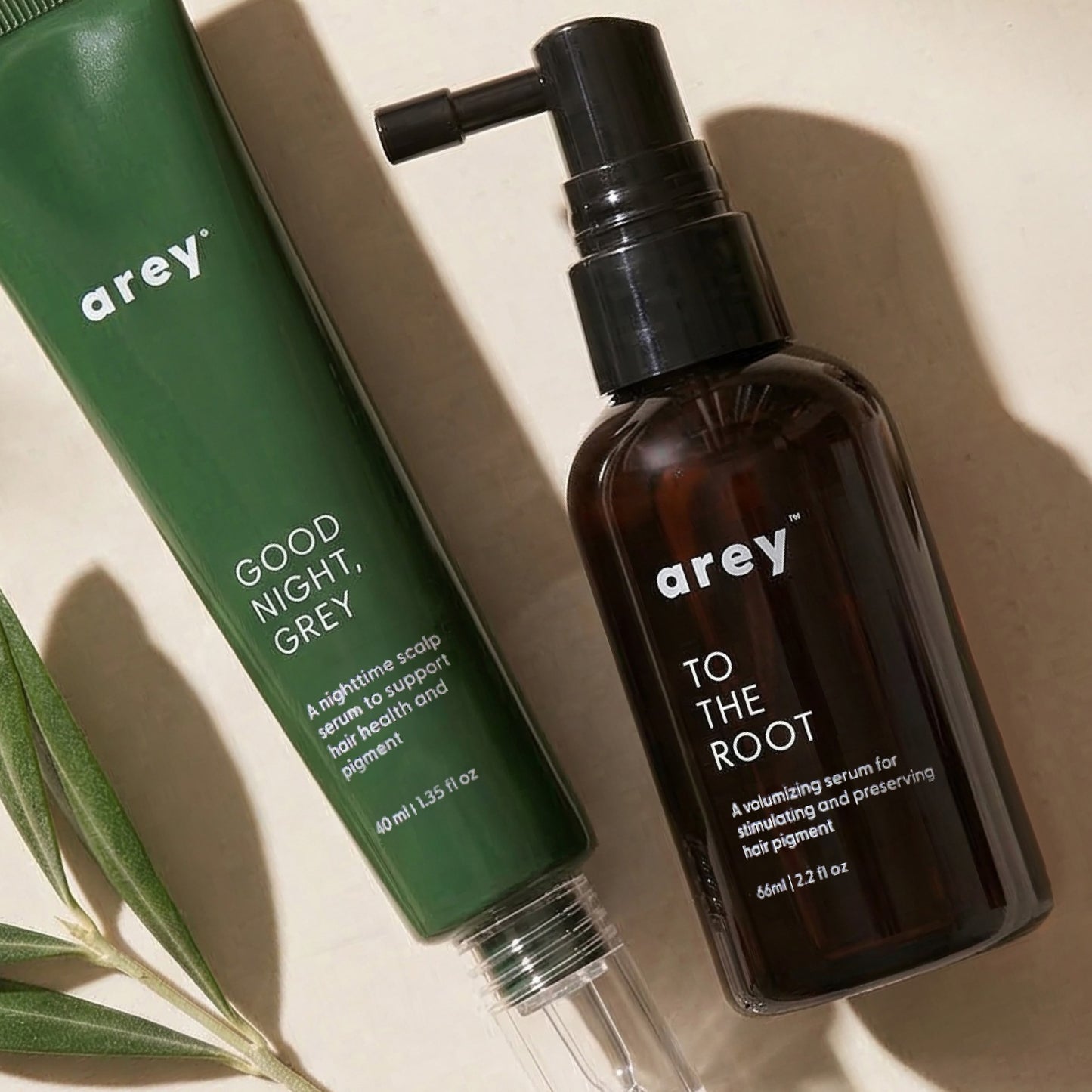 Two Arey products from The AM to PM Duo—a green tube and a brown pump bottle—are placed on a beige surface with a sprig of greenery nearby. Specially formulated for aging hair, this duo is designed to help reverse grey hair and promote thicker, fuller hair as part of your daily routine.