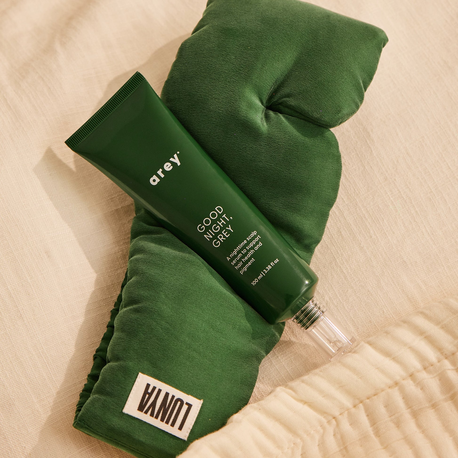 A green tube labeled Arey rests atop a dark green silk sleep mask with a white Lunya tag, both placed on a cream-colored fabric surface—perfectly introducing The Beauty Sleep Duo by Arey, an overnight scalp serum and mask set designed to support hair wellness.