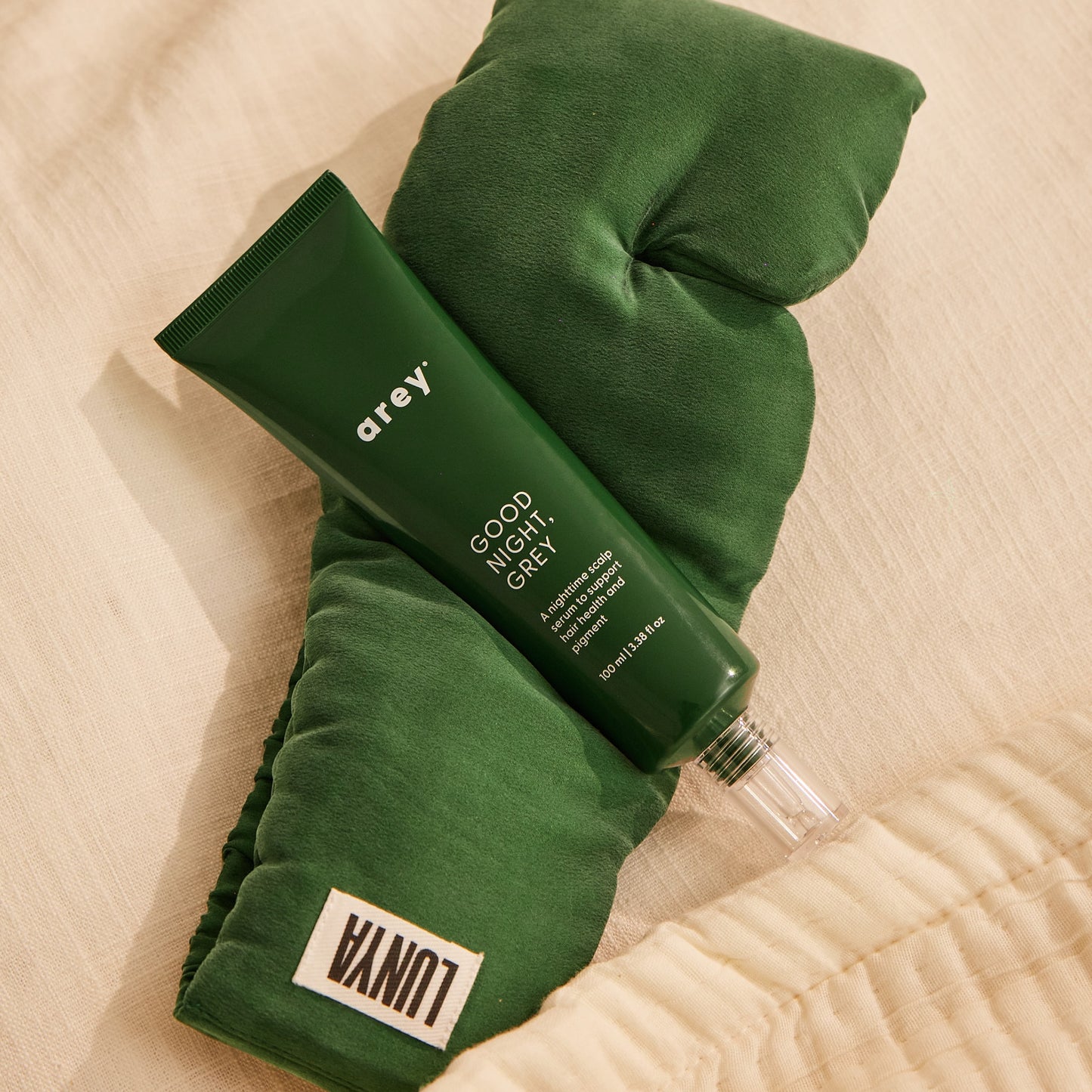 A green tube labeled Arey rests atop a dark green silk sleep mask with a white Lunya tag, both placed on a cream-colored fabric surface—perfectly introducing The Beauty Sleep Duo by Arey, an overnight scalp serum and mask set designed to support hair wellness.