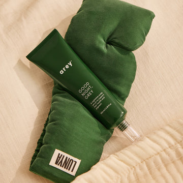 A green tube labeled Arey rests atop a dark green silk sleep mask with a white Lunya tag, both placed on a cream-colored fabric surface—perfectly introducing The Beauty Sleep Duo by Arey, an overnight scalp serum and mask set designed to support hair wellness.