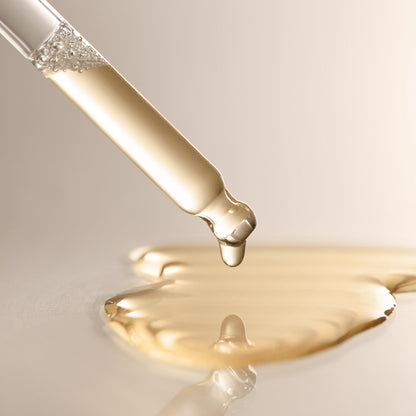 A close-up of a glass dropper releasing a drop of Arey’s Calm It Down Scalp Serum—a clear golden liquid—onto a smooth surface, with a small puddle forming below; expertly crafted to soothe and protect the scalp while defending against oxidative stress.