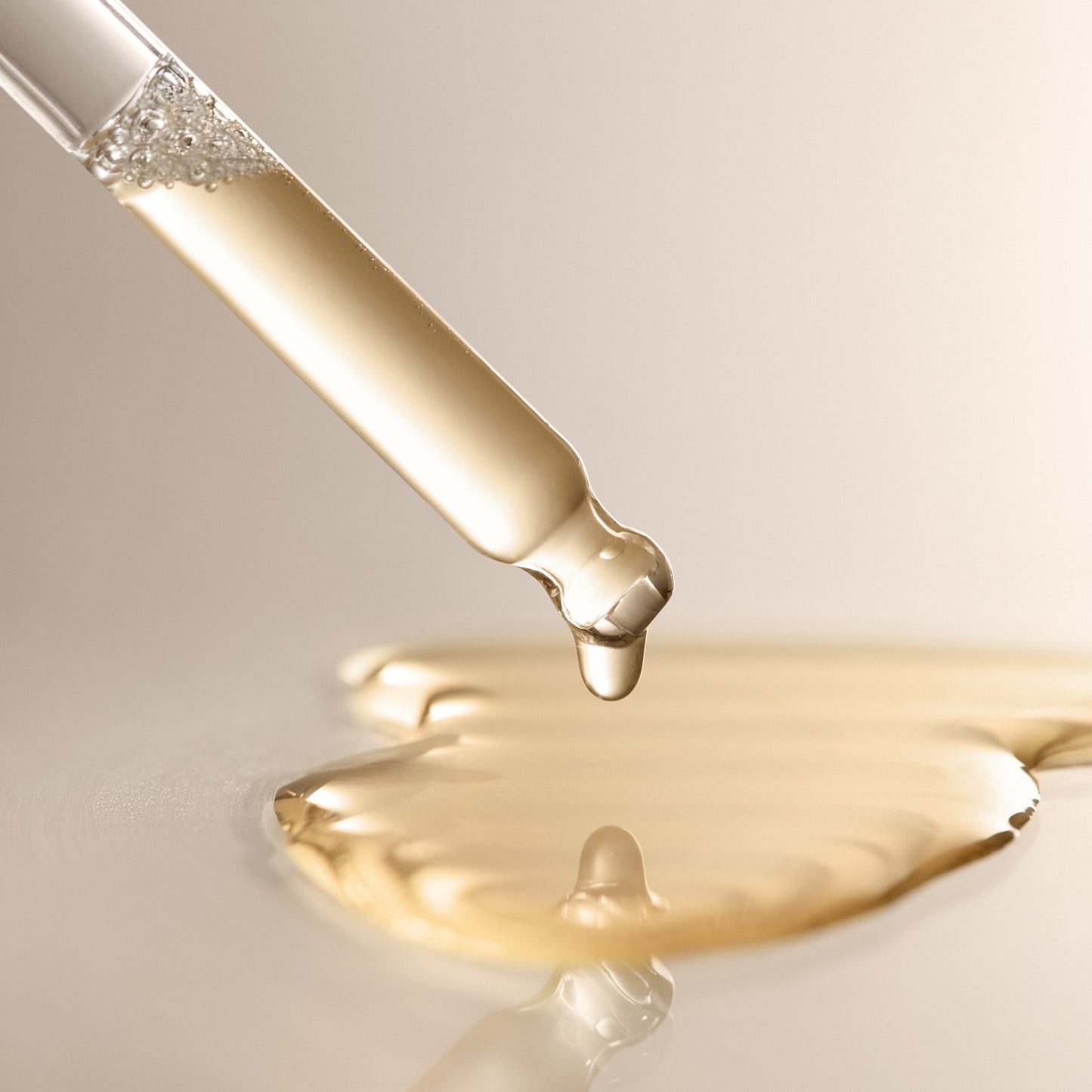 A close-up of a glass dropper releasing a drop of Arey’s Calm It Down Scalp Serum—a clear golden liquid—onto a smooth surface, with a small puddle forming below; expertly crafted to soothe and protect the scalp while defending against oxidative stress.