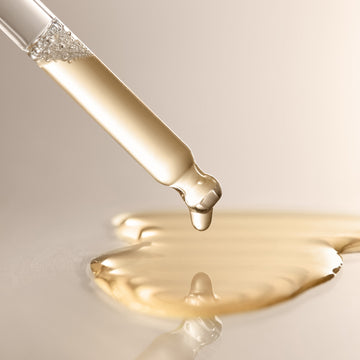 A close-up of a glass dropper releasing a drop of Arey’s Calm It Down Scalp Serum—a clear golden liquid—onto a smooth surface, with a small puddle forming below; expertly crafted to soothe and protect the scalp while defending against oxidative stress.