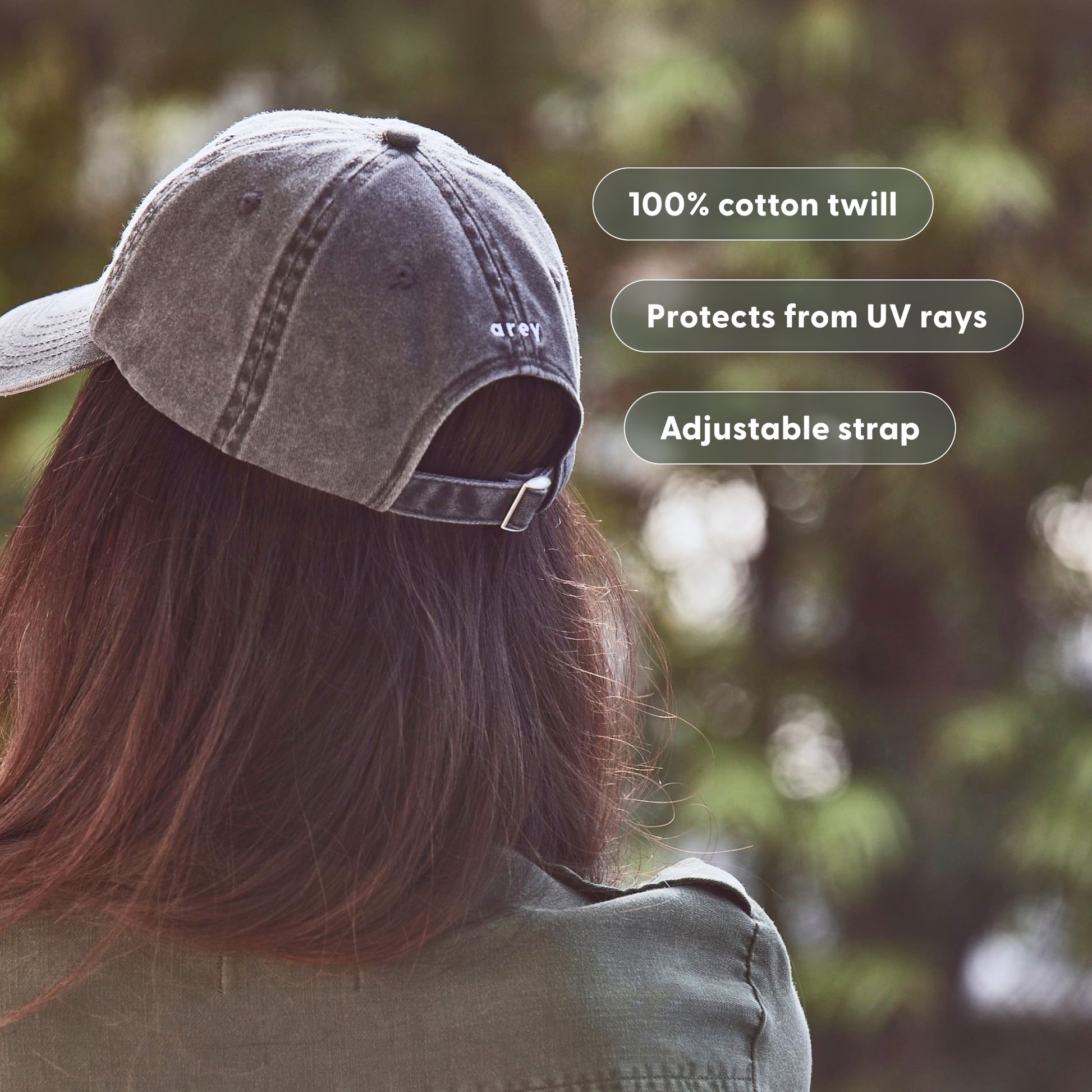 Lifestyle photo of a person wearing an arey branded cap outdoors, seen from behind with a green bokeh background. "arey" embroidered on the back of the cap. Feature badges overlay the image: 100% cotton twill, Protects from UV rays, Adjustable strap.