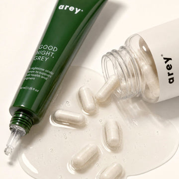 A green tube labeled Arey GOOD NIGHT and a clear bottle labeled Arey with white capsules spilling onto a glossy surface—The Dream Duo by Arey, specially formulated to help reduce greys and support melanin production for healthier-looking hair.