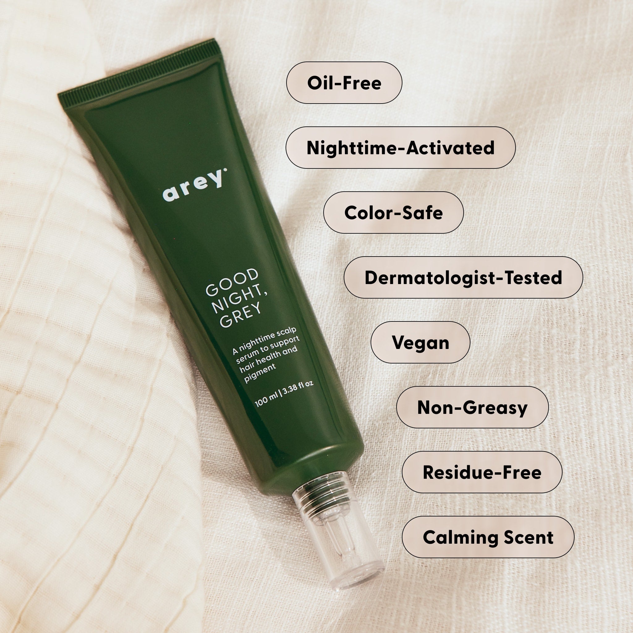 A green tube labeled Arey Good Night, Grey® lies on fabric, surrounded by labels highlighting its benefits: Oil-Free, Nighttime-Activated, Color-Safe, Dermatologist-Tested, Vegan, Non-Greasy, Residue-Free, and a Calming Scent. This overnight scalp treatment from Arey features hair growth peptides to nourish your scalp while you sleep.