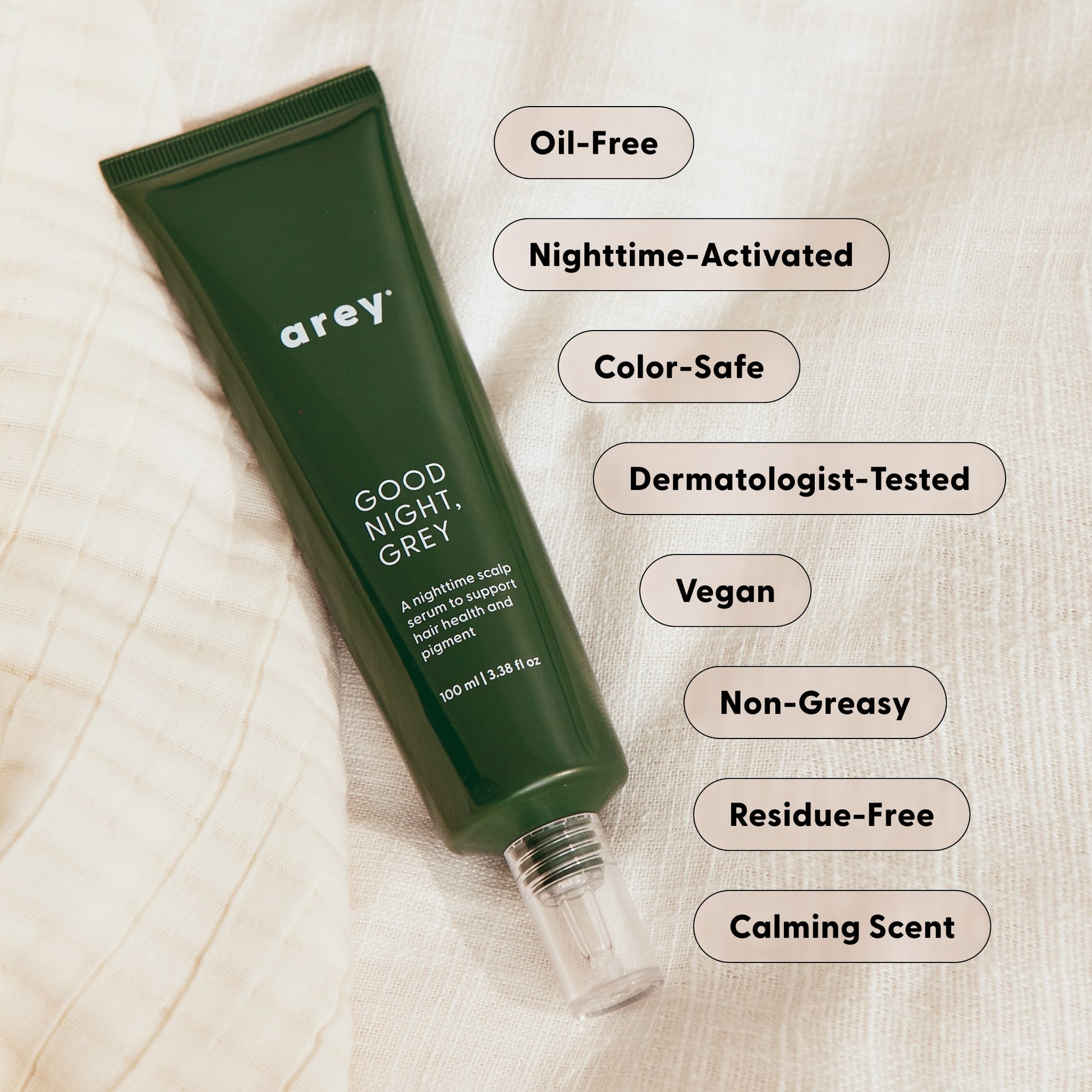 A green tube labeled Arey Good Night, Grey® lies on fabric, surrounded by labels highlighting its benefits: Oil-Free, Nighttime-Activated, Color-Safe, Dermatologist-Tested, Vegan, Non-Greasy, Residue-Free, and a Calming Scent. This overnight scalp treatment from Arey features hair growth peptides to nourish your scalp while you sleep.