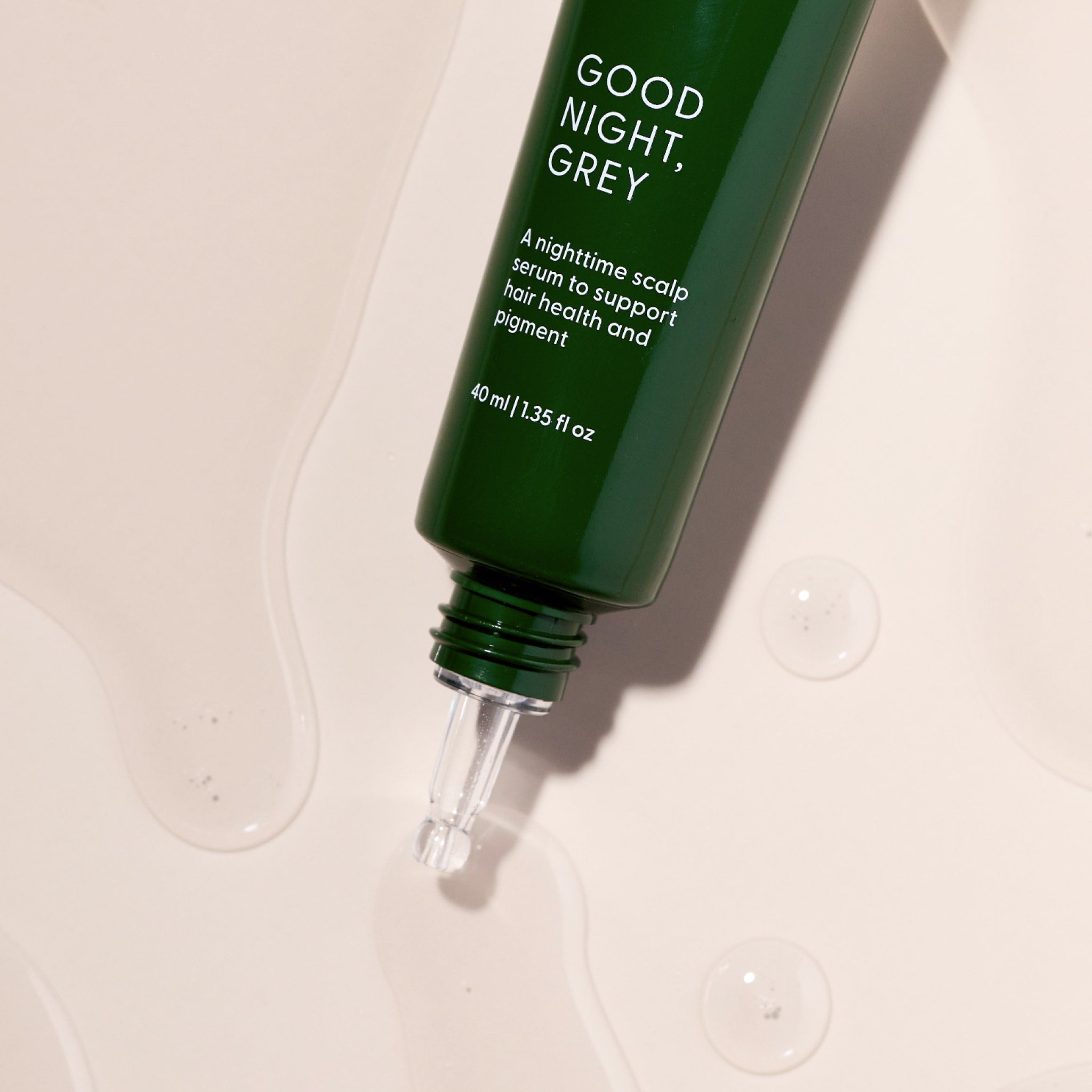A green bottle labeled Good Night, Grey® by Arey, featuring a dropper tip, rests on a light surface with clear liquid drops nearby. This overnight scalp treatment is specially formulated to support hair health and pigment, featuring hair growth peptides designed to promote fuller, more vibrant hair.