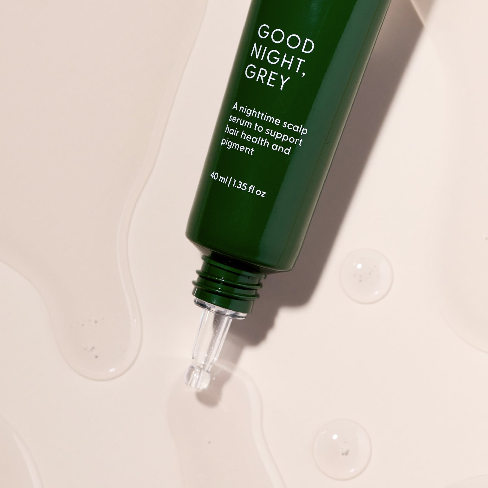 A green bottle labeled Good Night, Grey® by Arey, featuring a dropper tip, rests on a light surface with clear liquid drops nearby. This overnight scalp treatment is specially formulated to support hair health and pigment, featuring hair growth peptides designed to promote fuller, more vibrant hair.