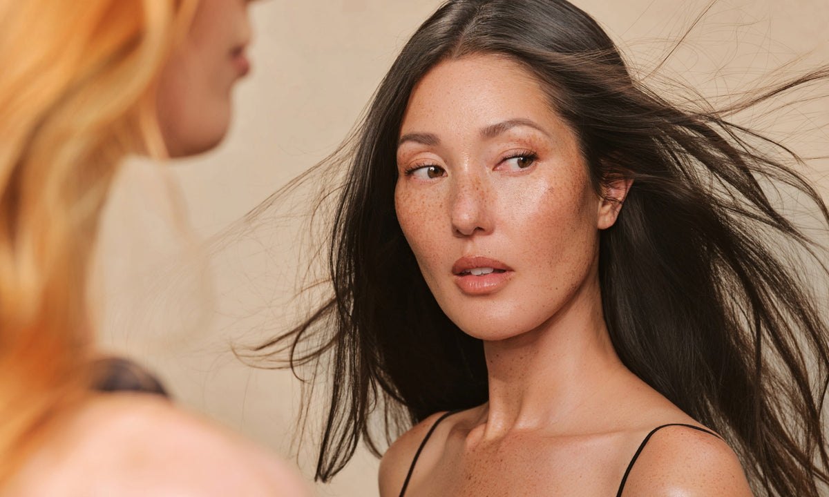 A person with long dark hair and visible freckles looks toward another person standing partially out of frame.