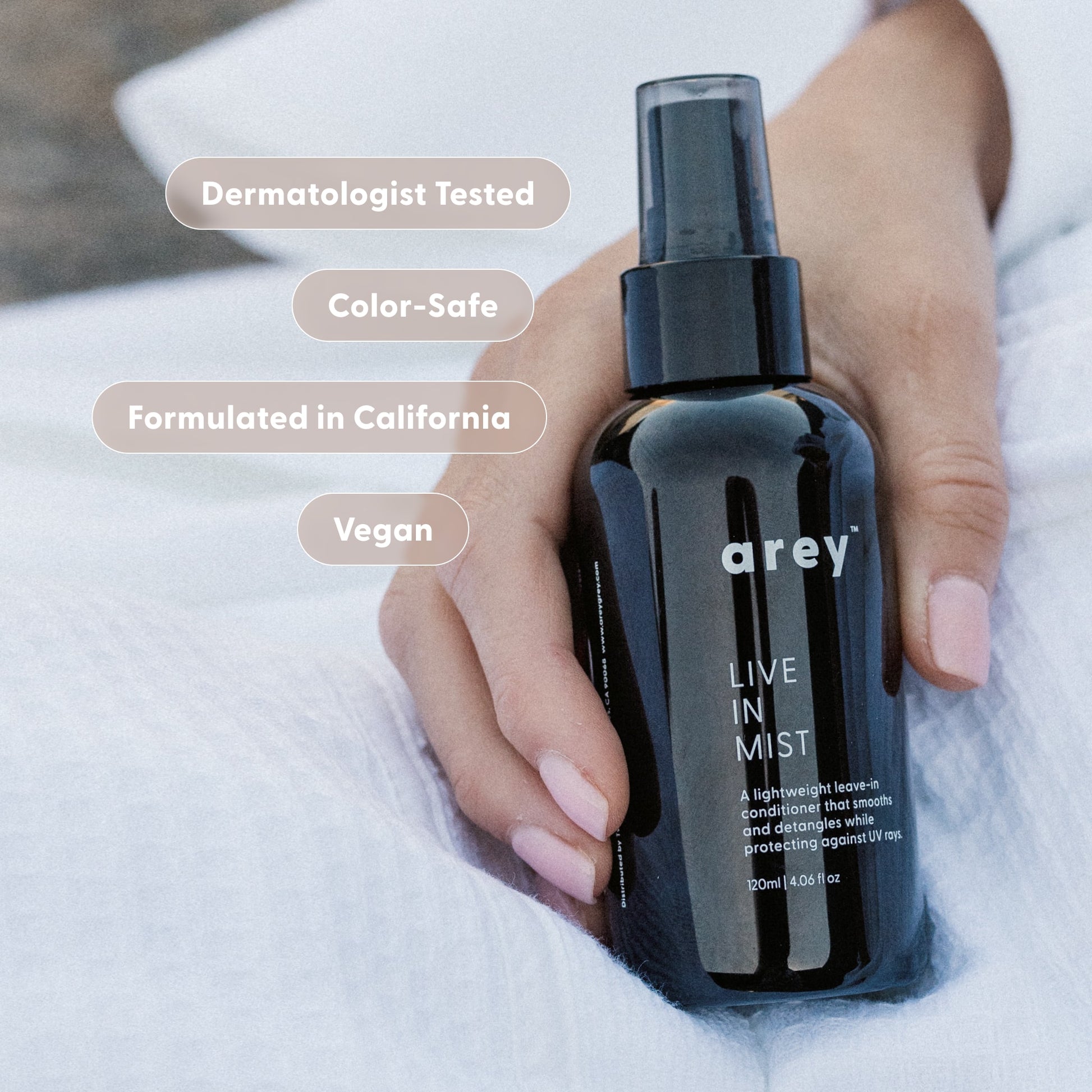 A hand holds a black bottle labeled Live In Mist Leave-In Conditioner by Arey on a white surface. Text bubbles read: Dermatologist Tested, Color-Safe, Formulated in California, Vegan, and enhanced with Raspberry Seed Oil for natural UV protection.