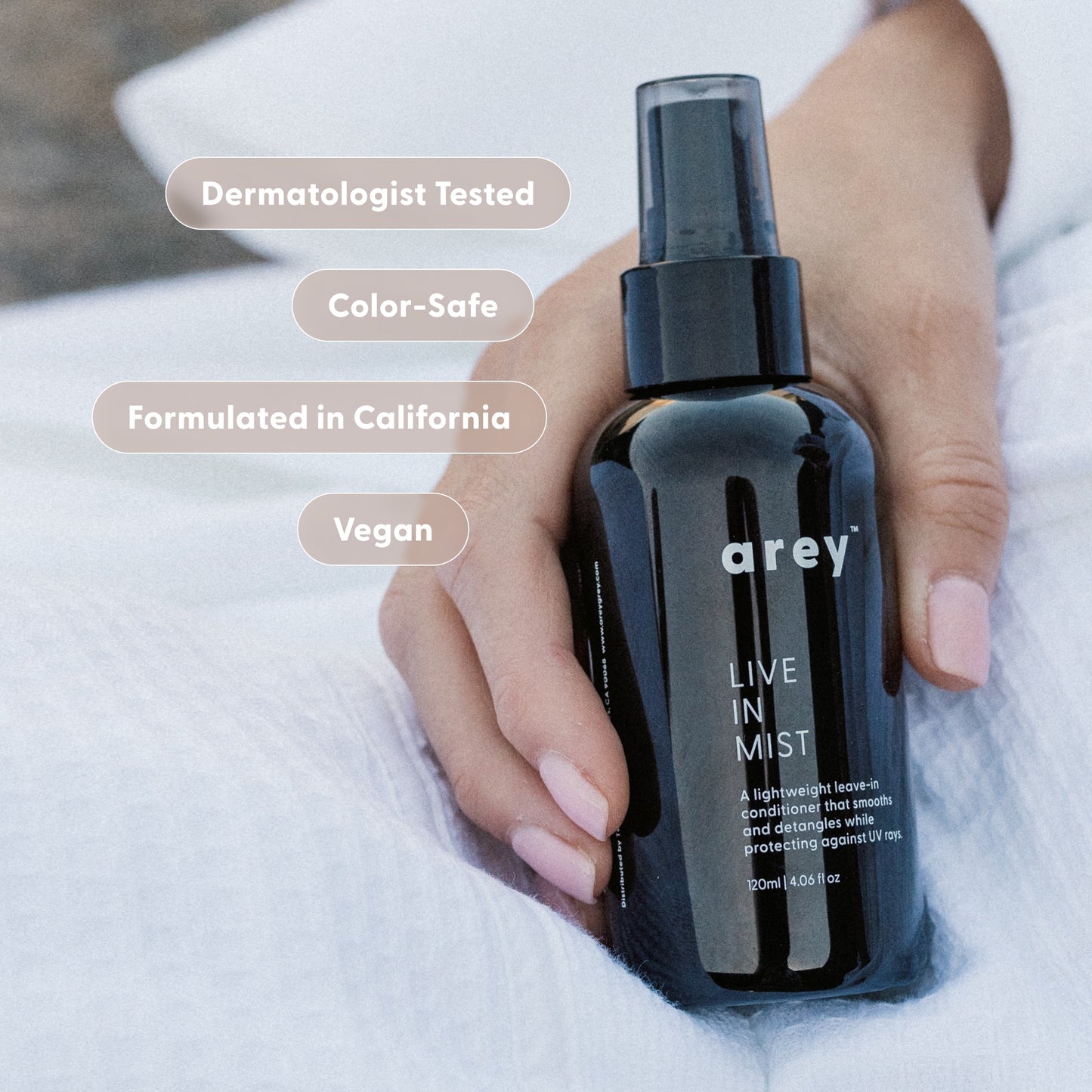 A hand holds a black bottle labeled Live In Mist Leave-In Conditioner by Arey on a white surface. Text bubbles read: Dermatologist Tested, Color-Safe, Formulated in California, Vegan, and enhanced with Raspberry Seed Oil for natural UV protection.