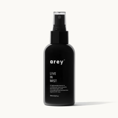 Product shot of arey Live In Mist dark glass spray bottle on a white/cream background. Label reads: arey™ LIVE IN MIST — "A lightweight leave-in conditioner that smooths and detangles with clinically proven protection against UV rays." 120ml | 4.06 fl oz.