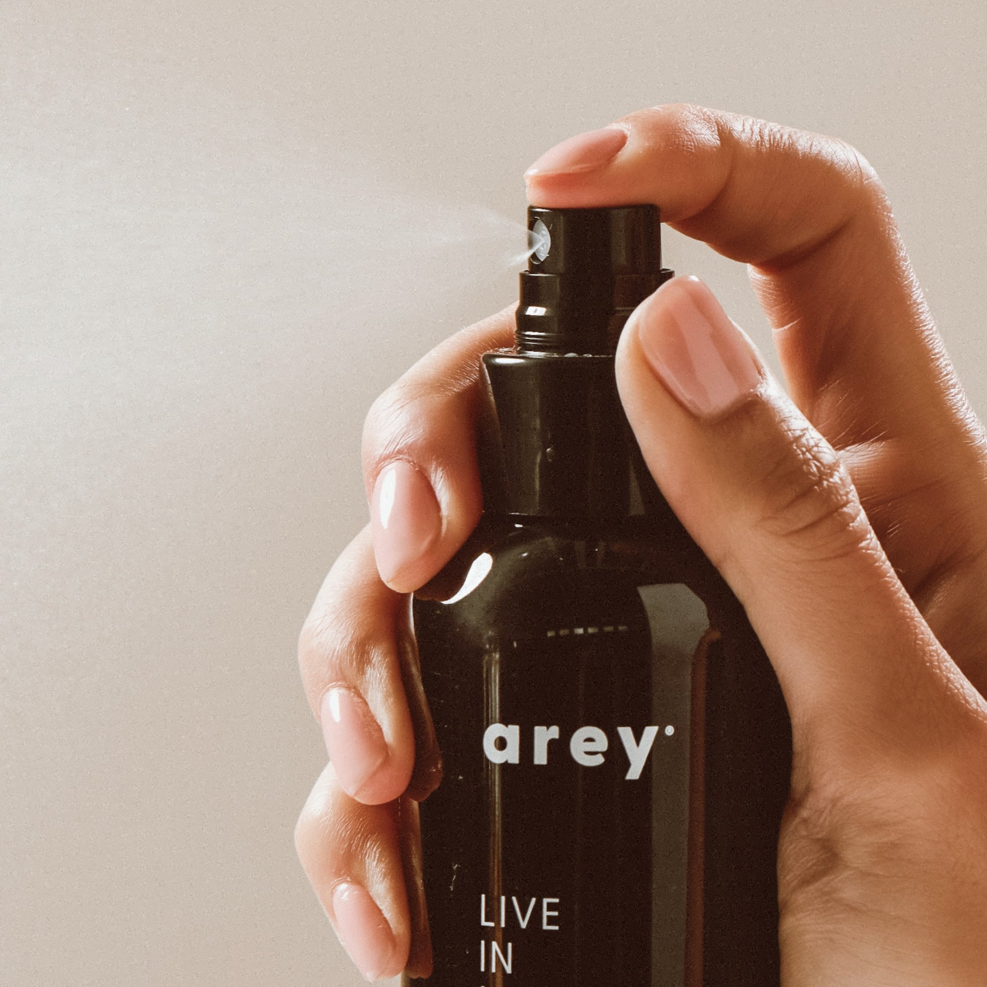 A hand holds a black spray bottle labeled “Arey Live In Mist Leave-In Conditioner,” pressing the nozzle to release a fine mist. The neutral background emphasizes the product and hand, showcasing its UV protection benefits and nourishment from Raspberry Seed Oil.