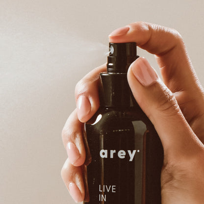 A hand holds a black spray bottle labeled “Arey Live In Mist Leave-In Conditioner,” pressing the nozzle to release a fine mist. The neutral background emphasizes the product and hand, showcasing its UV protection benefits and nourishment from Raspberry Seed Oil.