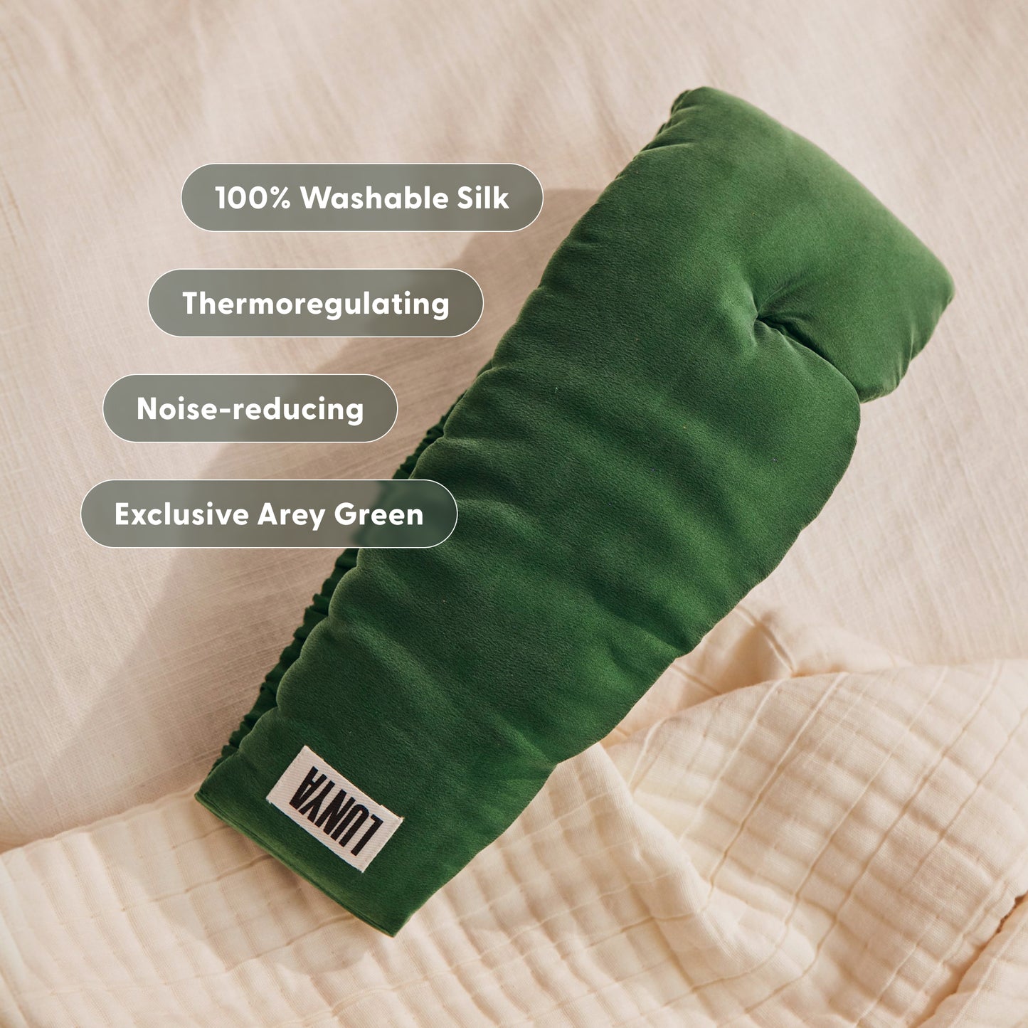 Lunya x Arey silk sleep mask in Exclusive Arey Green, resting on cream linen bedding. The mask features a LUNYA label. Feature badges overlay the image: 100% Washable Silk, Thermoregulating, Noise-reducing, Exclusive Arey Green.