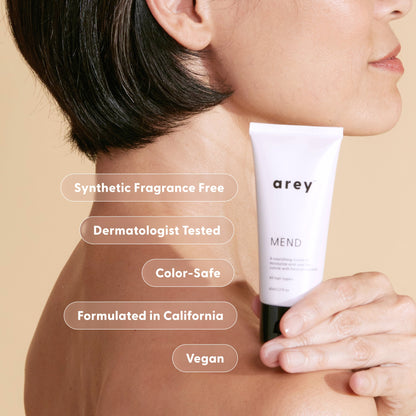 A person with short dark hair holds a tube labeled Arey Mend Heat Protectant Leave-In. Text highlights features: synthetic fragrance free, dermatologist tested, color-safe, formulated in California, and vegan. The product from Arey also helps reduce hair breakage and provides essential heat protection for healthier-looking hair.