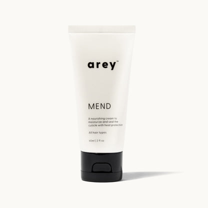 Product shot of Arey Mend hair treatment tube on a white/cream background. Label reads: arey® Mend. "A nourishing cream to moisturize and seal the cuticle with heat protection". All hair types. 60ml/2 fl oz.
