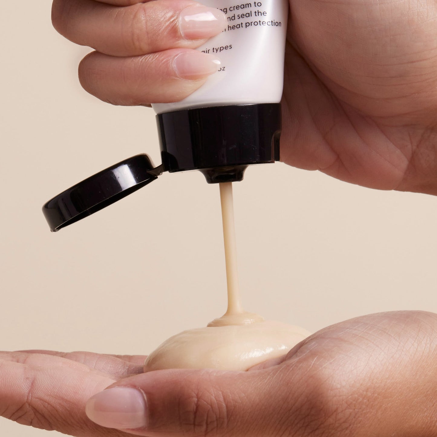 A person squeezes Arey’s Mend Heat Protectant Leave-In from a white bottle with a black flip cap onto the palm of their hand, against a plain beige background.