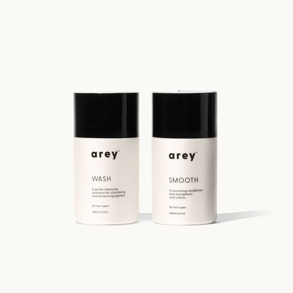 Two Arey Mini Duo travel-size bottles side by side on a white background. Left: arey® Wash — "A gentle cleansing shampoo for stimulating and preserving pigment", All hair types, 100ml/3.3 fl oz. Right: arey® Smooth — "A nourishing conditioner that strengthens and softens", All hair types, 100ml/3.3 fl oz.