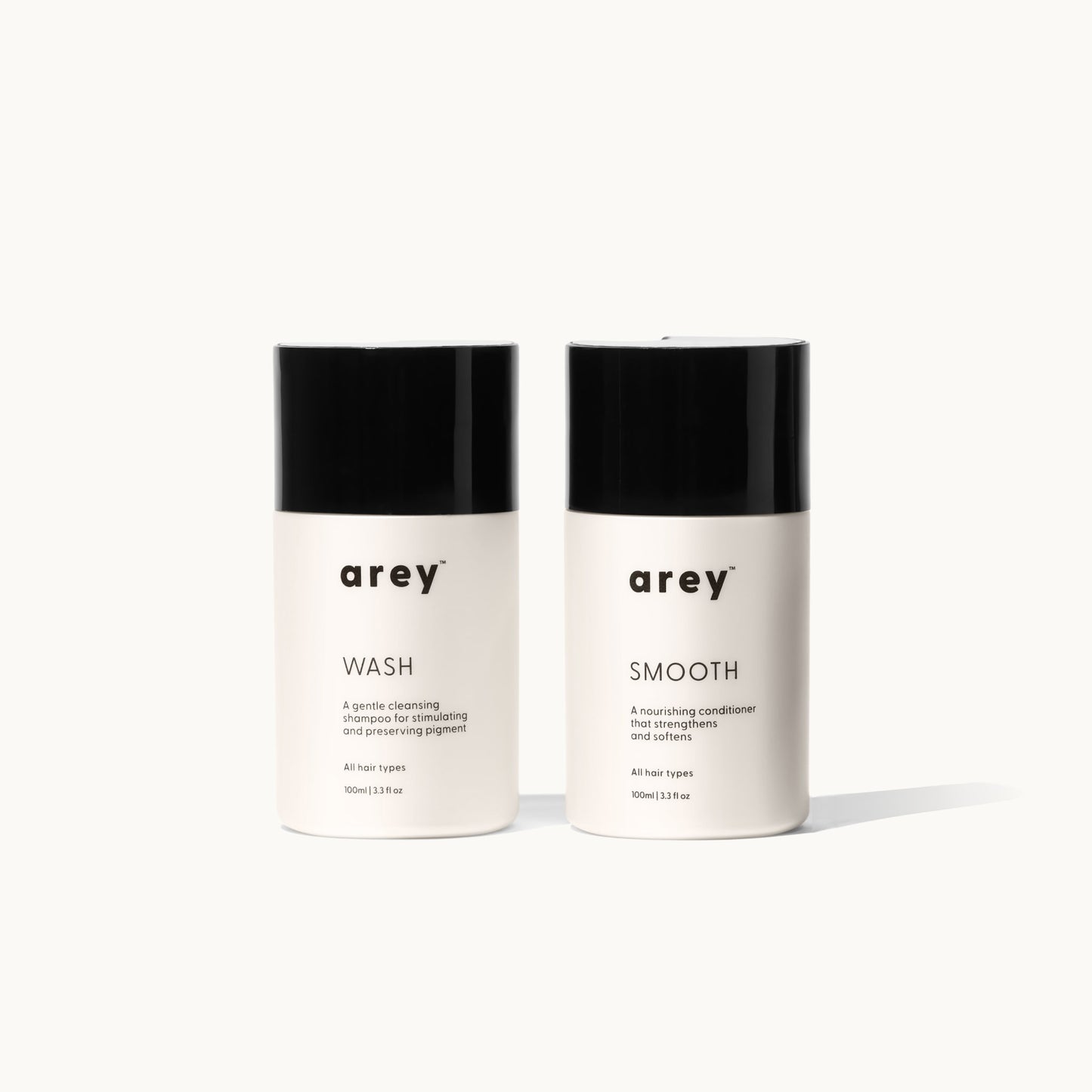Two Arey Mini Duo travel-size bottles side by side on a white background. Left: arey® Wash — "A gentle cleansing shampoo for stimulating and preserving pigment", All hair types, 100ml/3.3 fl oz. Right: arey® Smooth — "A nourishing conditioner that strengthens and softens", All hair types, 100ml/3.3 fl oz.