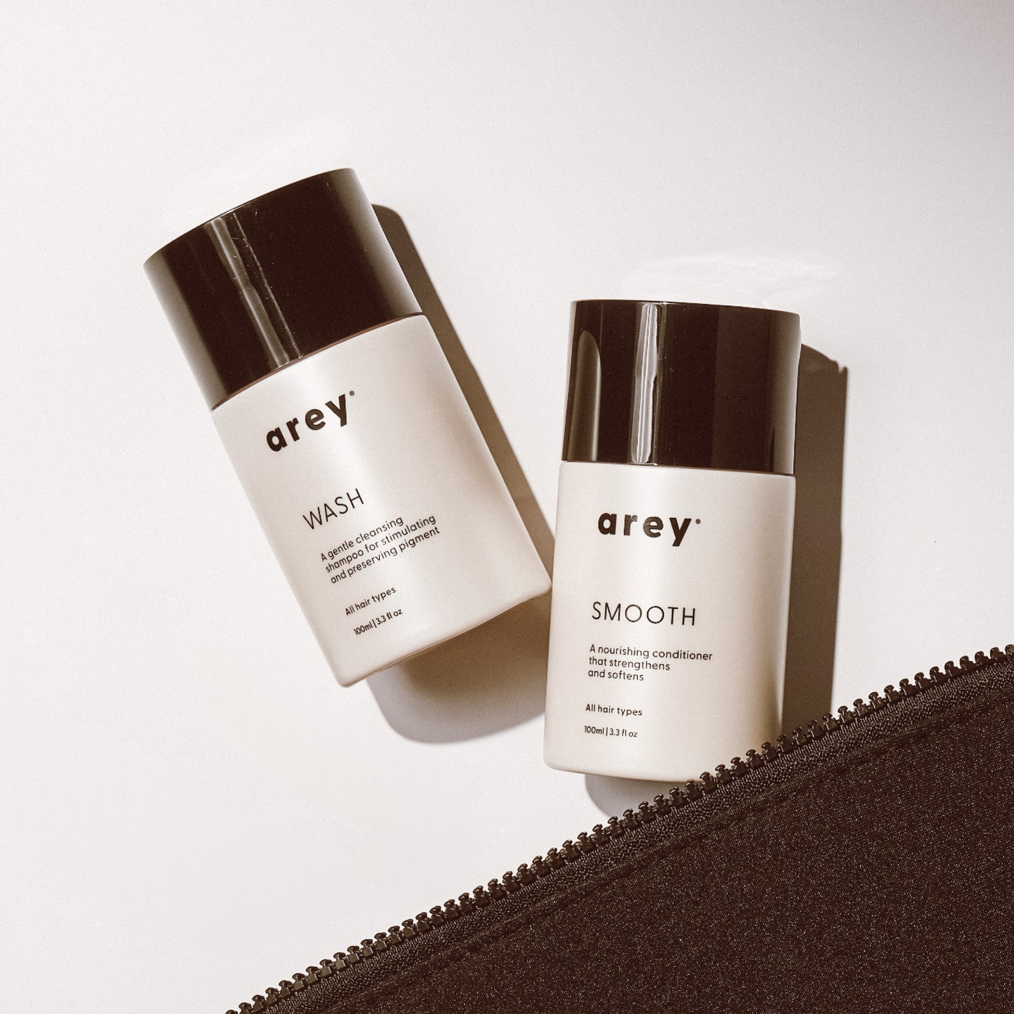Two Arey Mini Duo bottles, labeled WASH and SMOOTH, rest on a white surface beside a partially open black zipper pouch. Soft lighting creates gentle shadows, accentuating the minimal and modern design of these science-forward Arey hair care essentials made for healthier hair.