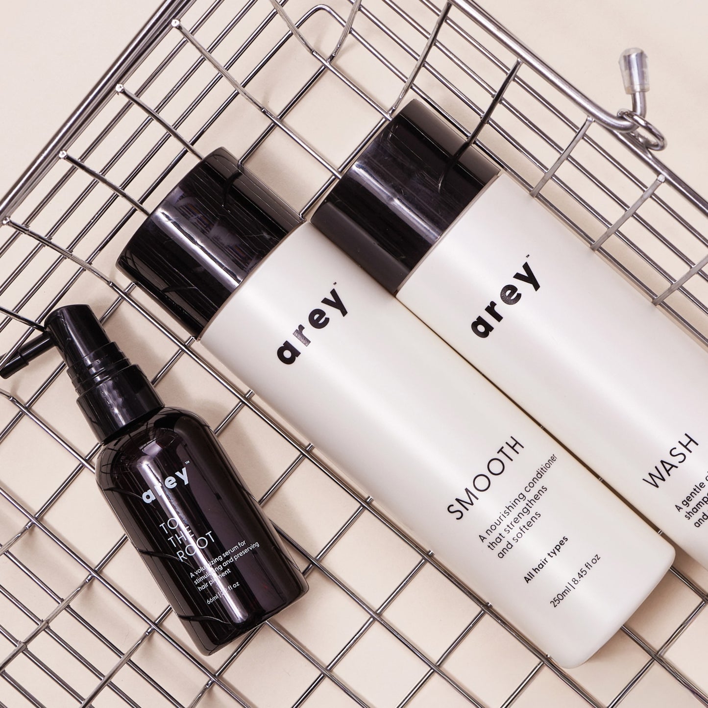 Three Arey haircare products—The Root Re-Set, Smooth, and Wash—are arranged diagonally on a metal wire tray against a beige background. The bottles feature sleek black and white minimalist labels, highlighting the brand’s commitment to scalp health and anti-aging hair care.