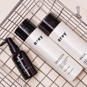 Three Arey haircare products—The Root Re-Set, Smooth, and Wash—are arranged diagonally on a metal wire tray against a beige background. The bottles feature sleek black and white minimalist labels, highlighting the brand’s commitment to scalp health and anti-aging hair care.