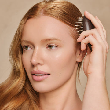 A woman with reddish brown hair using the Arey Scalp Brush, brushing through the side of her hair on a tan background.