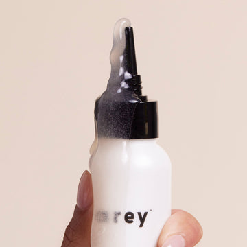 A hand holds a white bottle of Arey Scrub Scalp Exfoliant, featuring a black nozzle. Clear gel, likely containing pigment-preserving peptide and salicylic acid, overflows from the top. Part of the label is visible, displaying the letters “r e y” against a plain background.