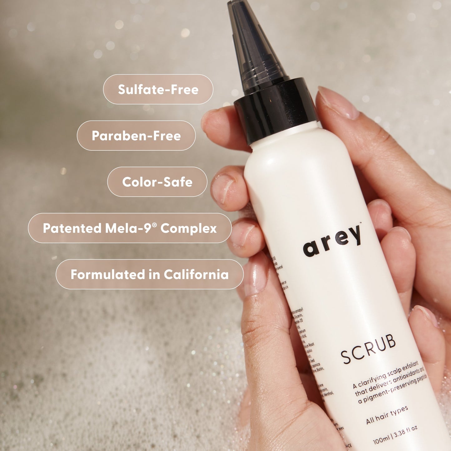 A hand holds a white Arey Scrub Scalp Exfoliant bottle above foamy water. Text bubbles read: Sulfate-Free, Paraben-Free, Color-Safe, Patented Mela-9® Complex with pigment-preserving peptide, and Formulated in California. Powered by salicylic acid for gentle scalp exfoliation.