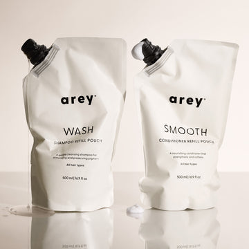 Arey Shower Duo Refill Pouches on a reflective surface. Left: Arey Wash Shampoo Refill Pouch (500ml / 16.9 fl oz) — A gentle cleansing shampoo for stimulating and preserving pigment. All hair types. Right: Arey Smooth Conditioner Refill Pouch (500ml / 16.9 fl oz) — A nourishing conditioner that strengthens and softens. All hair types.