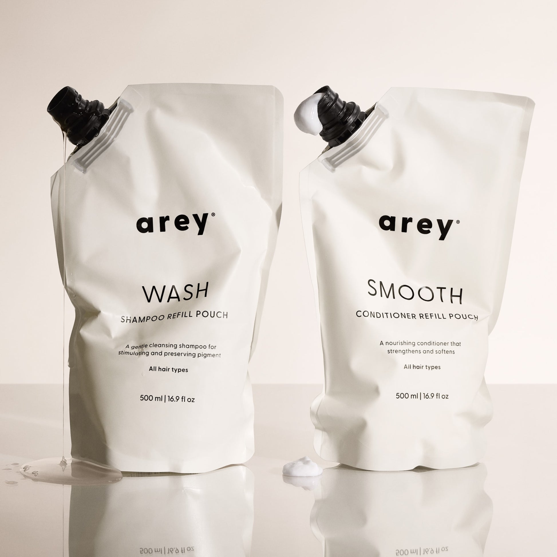 Two upright 500 ml white pouches labeled Arey The Shower Duo Refill feature minimalist black text, with a hint of product visible at the nozzle—showcasing essential in-shower care from Arey.