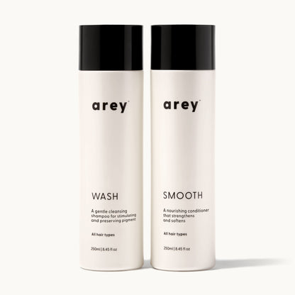 Arey Shower Duo on cream background. Two bottles side by side: Wash (A gentle cleansing shampoo for stimulating and preserving pigment. All hair types. 250ml / 8.45 fl oz) and Smooth (A nourishing conditioner that strengthens and softens. All hair types. 250ml / 8.45 fl oz).