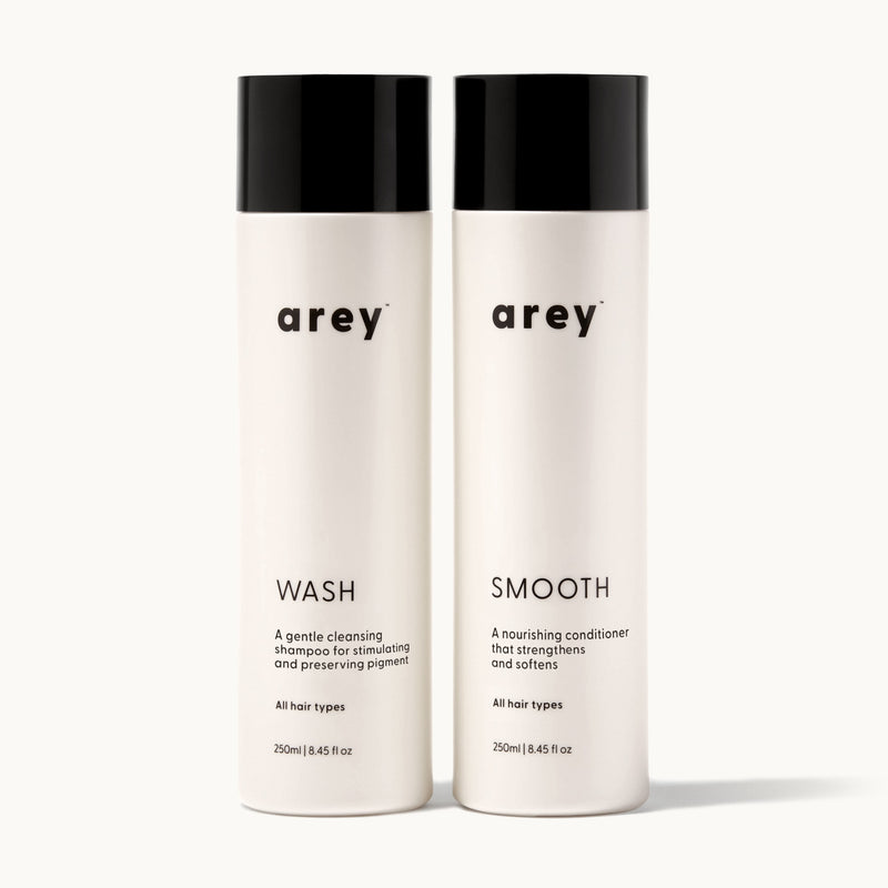 Arey Shower Duo on cream background. Two bottles side by side: Wash (A gentle cleansing shampoo for stimulating and preserving pigment. All hair types. 250ml / 8.45 fl oz) and Smooth (A nourishing conditioner that strengthens and softens. All hair types. 250ml / 8.45 fl oz).