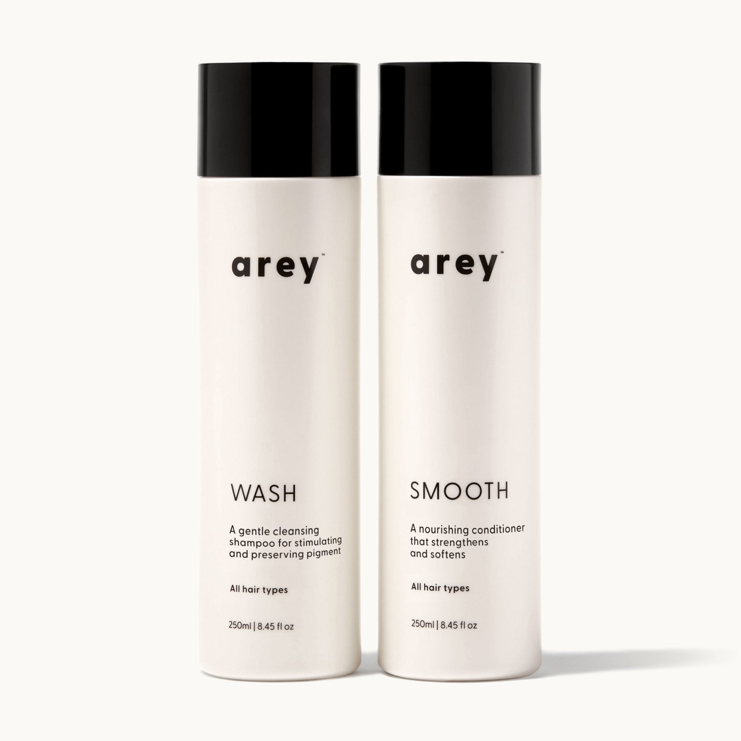 Arey Shower Duo on cream background. Two bottles side by side: Wash (A gentle cleansing shampoo for stimulating and preserving pigment. All hair types. 250ml / 8.45 fl oz) and Smooth (A nourishing conditioner that strengthens and softens. All hair types. 250ml / 8.45 fl oz).